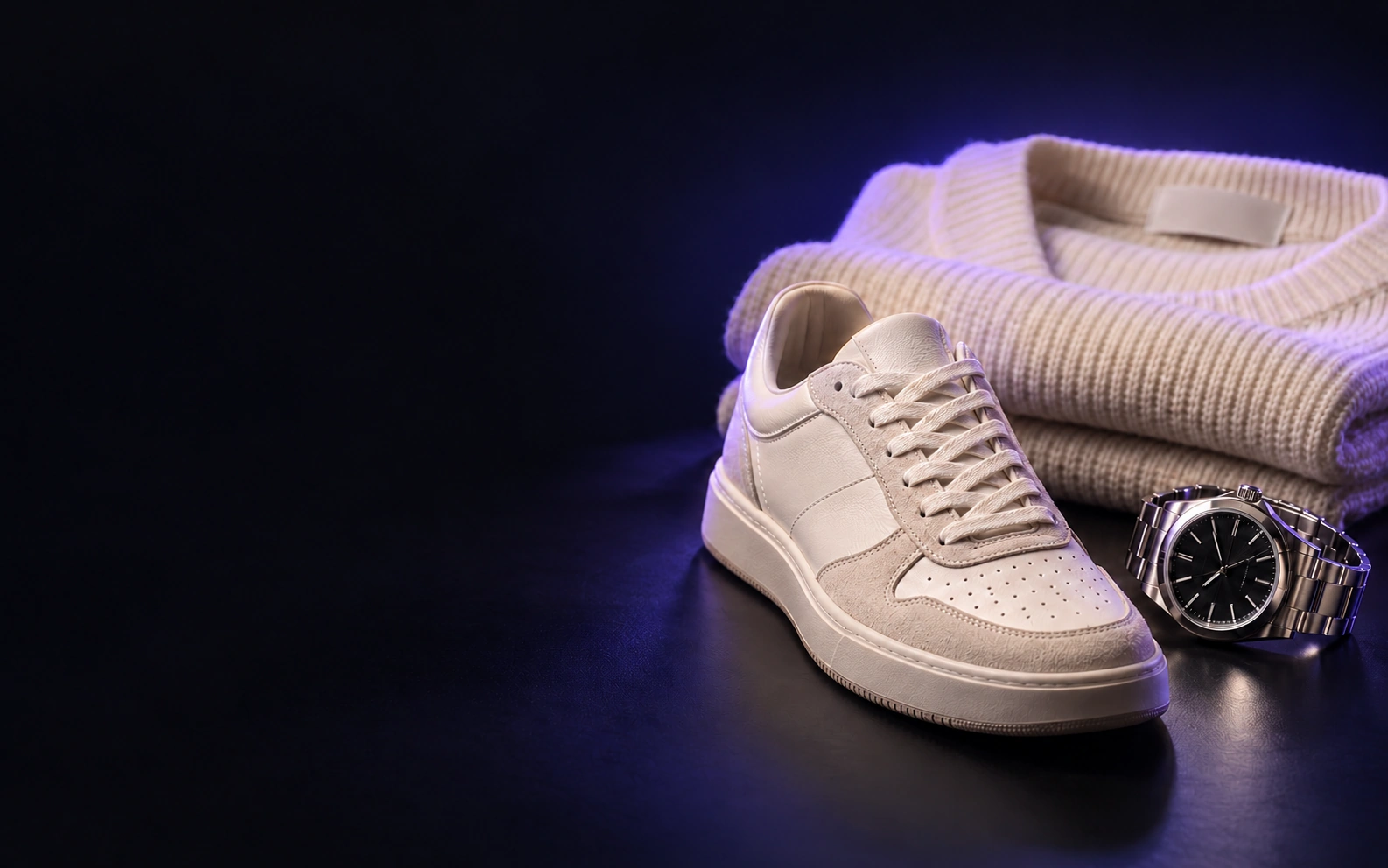 A cream sneaker, folded knitwear and a steel watch on a dark studio surface.
